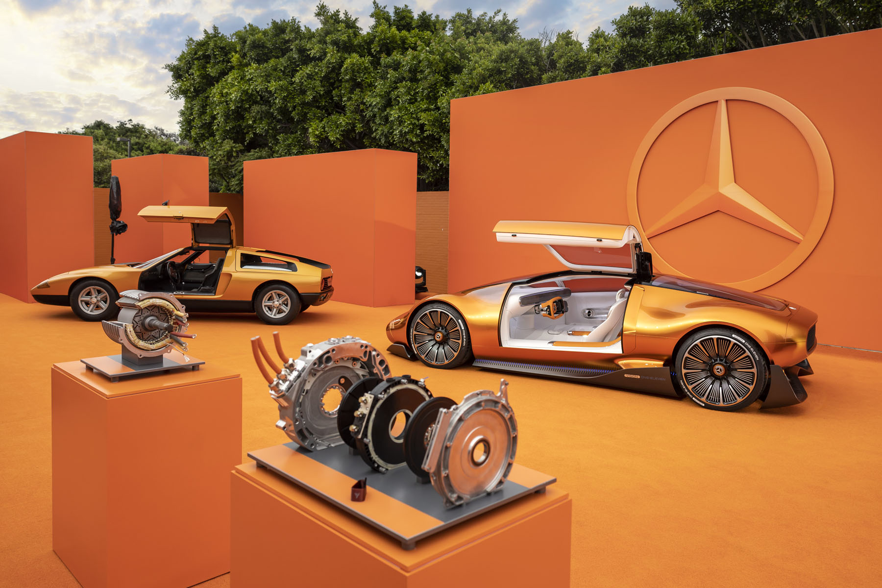 An exploded YASA axial flux motor showing its flat disc-shaped components, next to a conventional cylindrical radial motor, with the Mercedes-Benz C 111 and Vision One-Eleven in orange behind